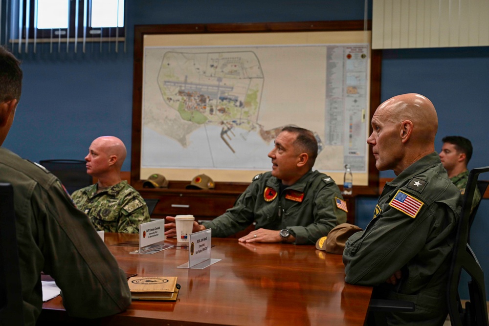 A military briefing or meeting is taking place in a conference room with several uniformed personnel seated around a wooden table. A large map is displayed on the wall behind them, appearing to show a coastal military installation or base. Personnel are wearing various U.S. military uniforms including flight suits and Navy working uniforms.