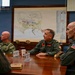A military briefing or meeting is taking place in a conference room with several uniformed personnel seated around a wooden table. A large map is displayed on the wall behind them, appearing to show a coastal military installation or base. Personnel are wearing various U.S. military uniforms including flight suits and Navy working uniforms.