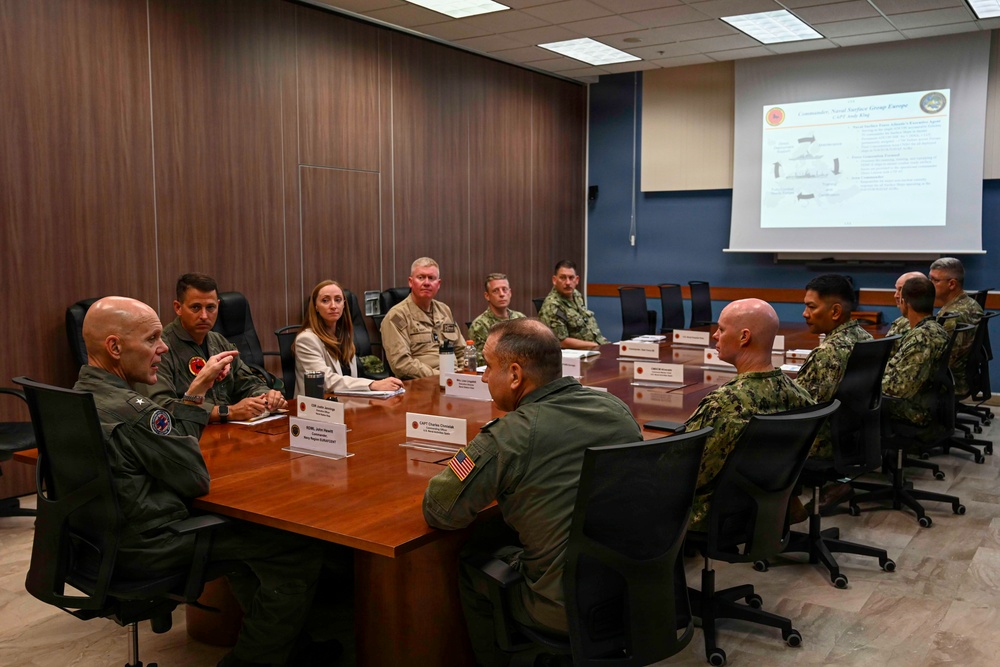 A formal military briefing is taking place in a conference room with approximately ten personnel seated around a large wooden table. The group includes uniformed military members from what appears to be the U.S. Navy and Marine Corps, along with at least one civilian attendee. A projected presentation slide is visible on the screen at the far end of the room, with text referencing 'Naval Surface Group Europe'.