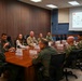 A formal military briefing is taking place in a conference room with approximately ten personnel seated around a large wooden table. The group includes uniformed military members from what appears to be the U.S. Navy and Marine Corps, along with at least one civilian attendee. A projected presentation slide is visible on the screen at the far end of the room, with text referencing 'Naval Surface Group Europe'.