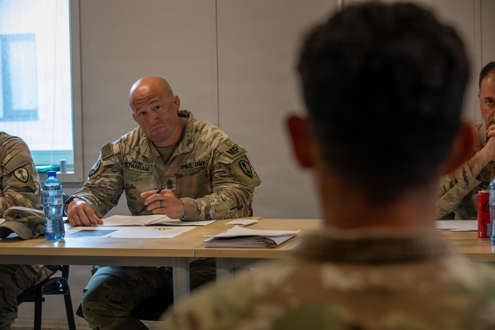 A U.S. Army Military Police (MP) soldier identified by name tape as 'PARHAM' sits at a table during what appears to be a briefing or meeting. He is wearing OCP (Operational Camouflage Pattern) uniform with U.S. Army and MP insignia, holding a pen and reviewing documents. The setting appears to be a military facility or forward operating base conference room.