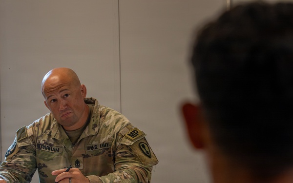 A U.S. Army Military Police (MP) soldier identified by name tape as 'PARHAM' sits at a table during what appears to be a briefing or meeting. He is wearing OCP (Operational Camouflage Pattern) uniform with U.S. Army and MP insignia, holding a pen and reviewing documents. The setting appears to be a military facility or forward operating base conference room.