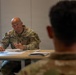 A U.S. Army Military Police (MP) soldier identified by name tape as 'PARHAM' sits at a table during what appears to be a briefing or meeting. He is wearing OCP (Operational Camouflage Pattern) uniform with U.S. Army and MP insignia, holding a pen and reviewing documents. The setting appears to be a military facility or forward operating base conference room.