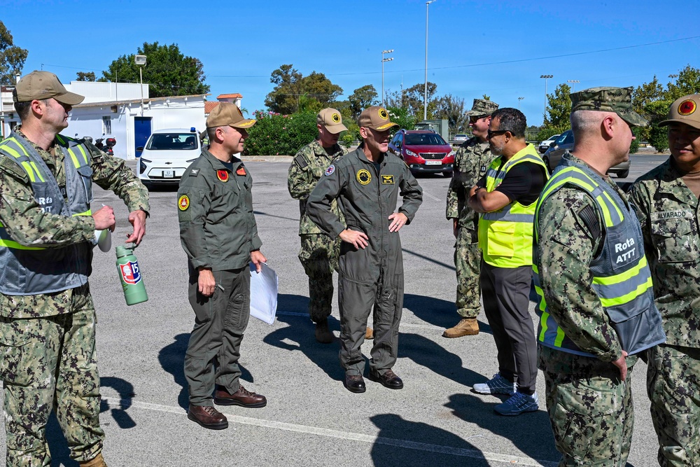 A group of military personnel are gathered in an outdoor parking lot area, appearing to be in a briefing or discussion formation. The group includes individuals in US Navy digital camouflage uniforms, olive/sage green flight suits, and high-visibility safety vests labeled 'Rota ATTT'. The setting appears to be Naval Station Rota, Spain, based on visible vest markings.