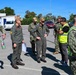 A group of military personnel are gathered in an outdoor parking lot area, appearing to be in a briefing or discussion formation. The group includes individuals in US Navy digital camouflage uniforms, olive/sage green flight suits, and high-visibility safety vests labeled 'Rota ATTT'. The setting appears to be Naval Station Rota, Spain, based on visible vest markings.