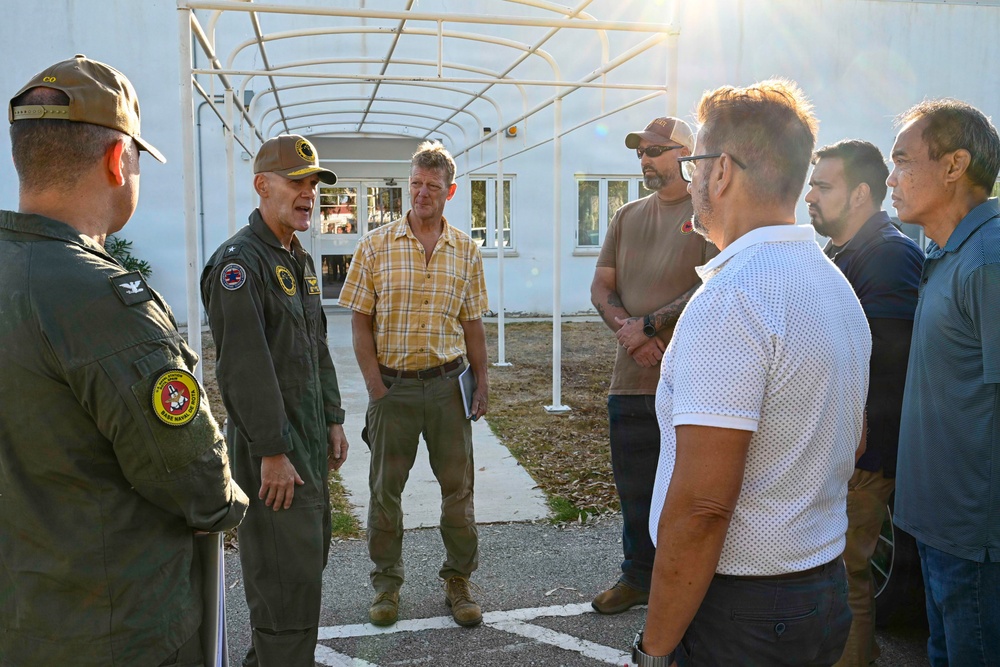 A group of approximately seven to eight individuals are gathered outdoors near a white building under a covered walkway structure. Two individuals on the left are wearing military flight suits with patches and caps, appearing to be in conversation with a mixed group of civilians. The setting appears to be a military installation or base facility.