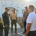 A group of approximately seven to eight individuals are gathered outdoors near a white building under a covered walkway structure. Two individuals on the left are wearing military flight suits with patches and caps, appearing to be in conversation with a mixed group of civilians. The setting appears to be a military installation or base facility.
