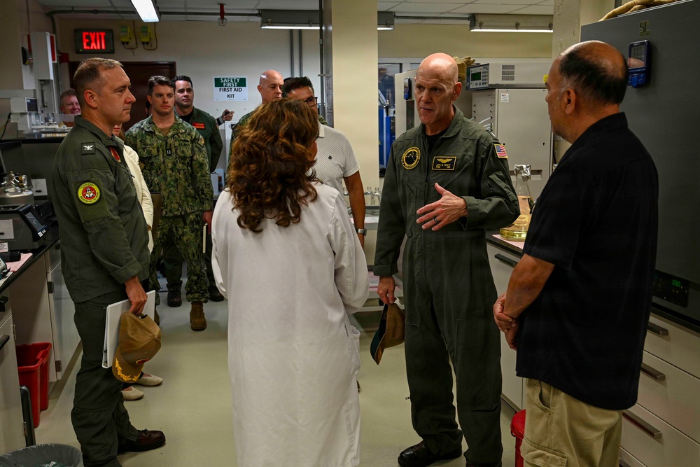A group of military personnel and civilians are gathered inside what appears to be a laboratory or research facility. A senior officer in an olive green flight suit is speaking to a person in a white lab coat and a civilian, while other uniformed personnel observe in the background. The setting suggests an official tour or briefing of a scientific or medical facility.
