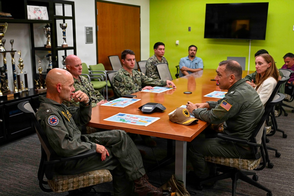 A group of military personnel and civilians are seated around a conference table in what appears to be a briefing or planning meeting. Several individuals are wearing green flight suits with patches, while others are in Navy camouflage uniforms. Documents with colorful maps or diagrams are spread on the table, suggesting an operational or logistical discussion.