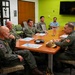 A group of military personnel and civilians are seated around a conference table in what appears to be a briefing or planning meeting. Several individuals are wearing green flight suits with patches, while others are in Navy camouflage uniforms. Documents with colorful maps or diagrams are spread on the table, suggesting an operational or logistical discussion.