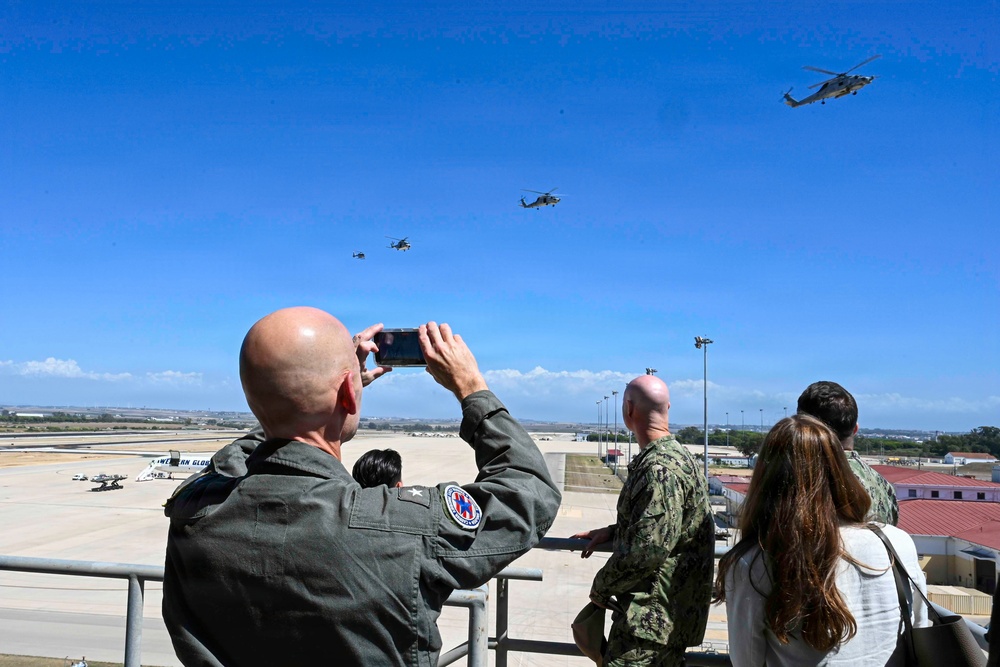 Military personnel and civilians observe a helicopter flyover from an elevated viewing platform at what appears to be a naval air station. A bald officer in olive green flight suit with a two-star insignia patch photographs the formation of helicopters using a smartphone. At least three helicopters are visible in the clear blue sky above the airfield.