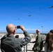 Military personnel and civilians observe a helicopter flyover from an elevated viewing platform at what appears to be a naval air station. A bald officer in olive green flight suit with a two-star insignia patch photographs the formation of helicopters using a smartphone. At least three helicopters are visible in the clear blue sky above the airfield.