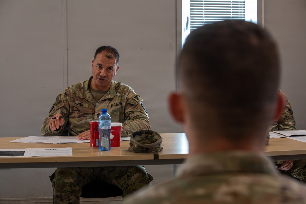A U.S. Army soldier named 'Smith' is seated at a table appearing to conduct or participate in a briefing or meeting. He is speaking and gesturing with his hand toward other military personnel seated across from him. The setting appears to be an indoor military facility or conference room.