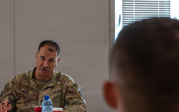 A U.S. Army soldier named 'Smith' is seated at a table appearing to conduct or participate in a briefing or meeting. He is speaking and gesturing with his hand toward other military personnel seated across from him. The setting appears to be an indoor military facility or conference room.