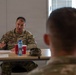 A U.S. Army soldier named 'Smith' is seated at a table appearing to conduct or participate in a briefing or meeting. He is speaking and gesturing with his hand toward other military personnel seated across from him. The setting appears to be an indoor military facility or conference room.