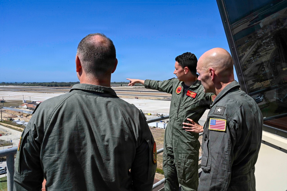 Three military personnel in olive/sage green flight suits are standing on what appears to be an elevated observation deck or control tower, overlooking a large airfield or military base. One individual in the center is pointing toward the runway area, appearing to brief or orient the others. The person on the right displays a one-star general insignia and a US flag patch on their sleeve.