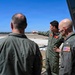 Three military personnel in olive/sage green flight suits are standing on what appears to be an elevated observation deck or control tower, overlooking a large airfield or military base. One individual in the center is pointing toward the runway area, appearing to brief or orient the others. The person on the right displays a one-star general insignia and a US flag patch on their sleeve.