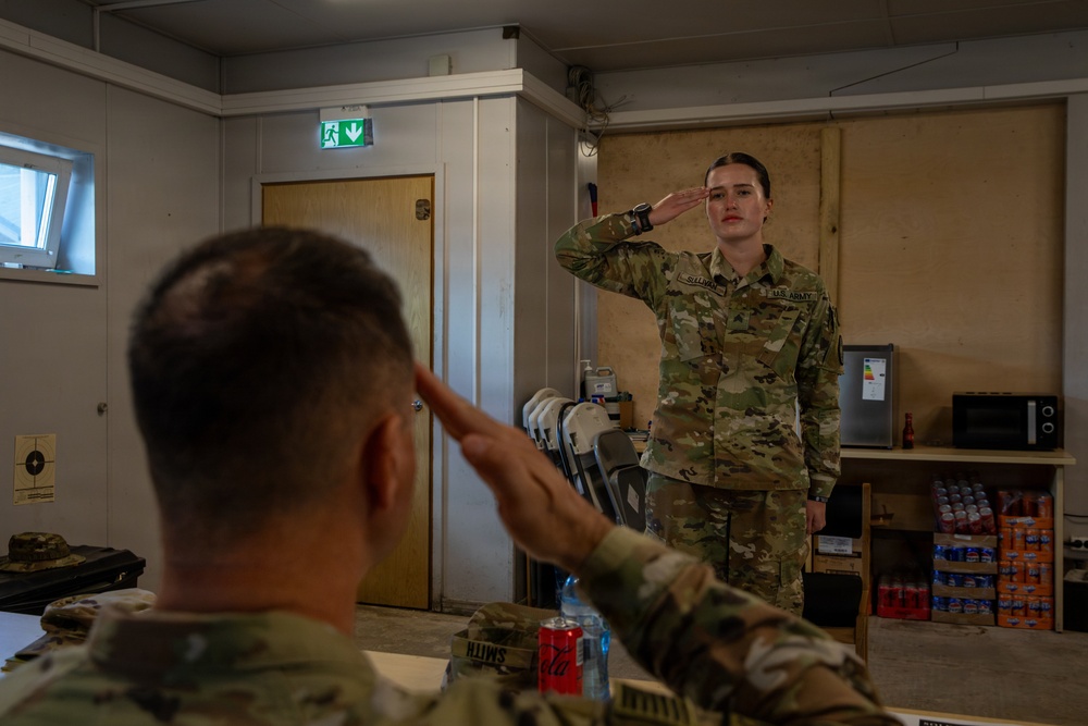 Two U.S. Army soldiers are exchanging salutes inside what appears to be a military facility or barracks room. The soldier in the foreground (facing away, name tape reading 'SMITH') is seated at a table and returning a salute to a standing female soldier whose name tape reads 'SULLIVAN'. The setting suggests a formal military interaction, possibly a report or briefing scenario.