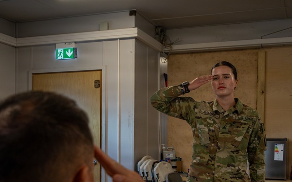 Two U.S. Army soldiers are exchanging salutes inside what appears to be a military facility or barracks room. The soldier in the foreground (facing away, name tape reading 'SMITH') is seated at a table and returning a salute to a standing female soldier whose name tape reads 'SULLIVAN'. The setting suggests a formal military interaction, possibly a report or briefing scenario.