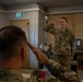 Two U.S. Army soldiers are exchanging salutes inside what appears to be a military facility or barracks room. The soldier in the foreground (facing away, name tape reading 'SMITH') is seated at a table and returning a salute to a standing female soldier whose name tape reads 'SULLIVAN'. The setting suggests a formal military interaction, possibly a report or briefing scenario.
