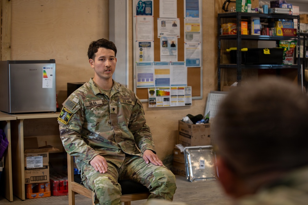 A U.S. Army soldier named Silva, wearing Operational Camouflage Pattern (OCP) uniform, is seated on a wooden chair and appears to be speaking or briefing to at least one other person visible in the foreground. The setting appears to be an informal common room or break area on a military installation, with a bulletin board, shelving unit with supplies, and a small refrigerator visible in the background. The atmosphere suggests a casual briefing, counseling session, or informal discussion.