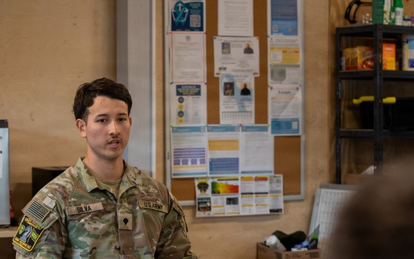 A U.S. Army soldier named Silva, wearing Operational Camouflage Pattern (OCP) uniform, is seated on a wooden chair and appears to be speaking or briefing to at least one other person visible in the foreground. The setting appears to be an informal common room or break area on a military installation, with a bulletin board, shelving unit with supplies, and a small refrigerator visible in the background. The atmosphere suggests a casual briefing, counseling session, or informal discussion.