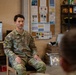 A U.S. Army soldier named Silva, wearing Operational Camouflage Pattern (OCP) uniform, is seated on a wooden chair and appears to be speaking or briefing to at least one other person visible in the foreground. The setting appears to be an informal common room or break area on a military installation, with a bulletin board, shelving unit with supplies, and a small refrigerator visible in the background. The atmosphere suggests a casual briefing, counseling session, or informal discussion.