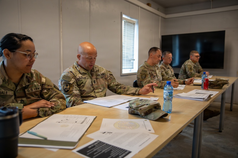 Five U.S. Army soldiers in MultiCam/OCP camouflage uniforms are seated at tables in what appears to be a briefing or meeting room. The soldiers are reviewing documents and engaged in discussion, with one soldier gesturing while speaking. The setting appears to be a temporary or forward operating facility based on the modular wall construction.