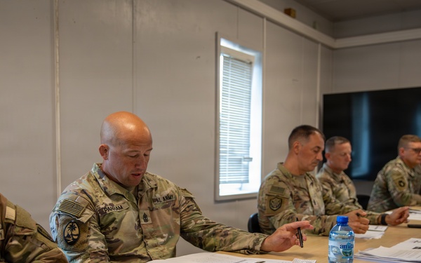 Five U.S. Army soldiers in MultiCam/OCP camouflage uniforms are seated at tables in what appears to be a briefing or meeting room. The soldiers are reviewing documents and engaged in discussion, with one soldier gesturing while speaking. The setting appears to be a temporary or forward operating facility based on the modular wall construction.