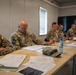Five U.S. Army soldiers in MultiCam/OCP camouflage uniforms are seated at tables in what appears to be a briefing or meeting room. The soldiers are reviewing documents and engaged in discussion, with one soldier gesturing while speaking. The setting appears to be a temporary or forward operating facility based on the modular wall construction.
