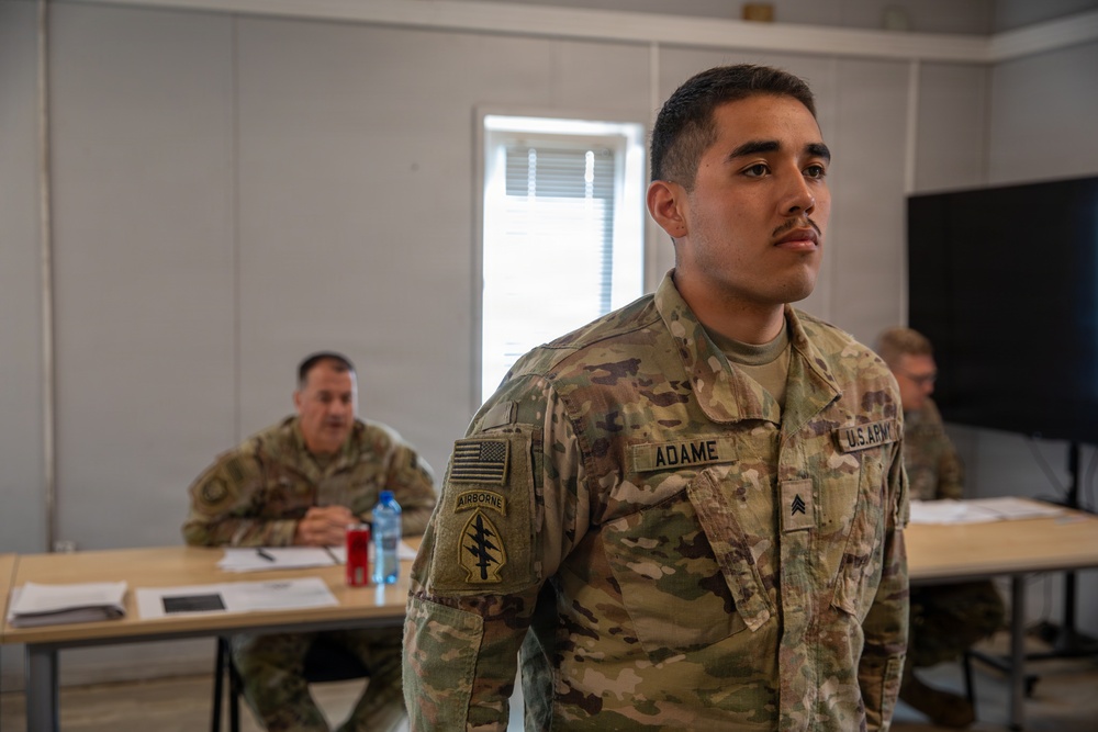 A U.S. Army Sergeant named Adame stands at attention in what appears to be a classroom or briefing room setting. He is wearing OCP (Operational Camouflage Pattern) uniform with an Airborne tab and Special Forces patch visible on his left shoulder. Two other soldiers are seated at tables in the background.