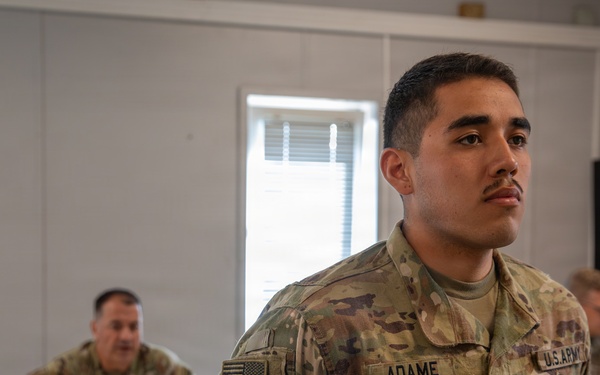 A U.S. Army Sergeant named Adame stands at attention in what appears to be a classroom or briefing room setting. He is wearing OCP (Operational Camouflage Pattern) uniform with an Airborne tab and Special Forces patch visible on his left shoulder. Two other soldiers are seated at tables in the background.