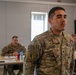 A U.S. Army Sergeant named Adame stands at attention in what appears to be a classroom or briefing room setting. He is wearing OCP (Operational Camouflage Pattern) uniform with an Airborne tab and Special Forces patch visible on his left shoulder. Two other soldiers are seated at tables in the background.