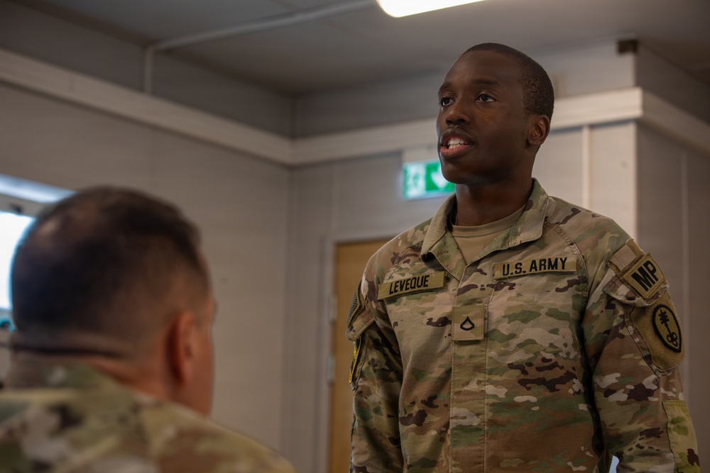 A U.S. Army Military Police soldier named Leveque, wearing OCP (Operational Camouflage Pattern) uniform, is engaged in conversation with another soldier whose back is to the camera. The setting appears to be an indoor military facility, possibly in Europe based on the green emergency exit sign visible in the background. The soldier appears to be speaking or responding to the other individual.