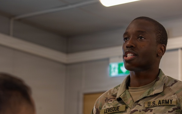A U.S. Army Military Police soldier named Leveque, wearing OCP (Operational Camouflage Pattern) uniform, is engaged in conversation with another soldier whose back is to the camera. The setting appears to be an indoor military facility, possibly in Europe based on the green emergency exit sign visible in the background. The soldier appears to be speaking or responding to the other individual.