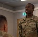 A U.S. Army Military Police soldier named Leveque, wearing OCP (Operational Camouflage Pattern) uniform, is engaged in conversation with another soldier whose back is to the camera. The setting appears to be an indoor military facility, possibly in Europe based on the green emergency exit sign visible in the background. The soldier appears to be speaking or responding to the other individual.