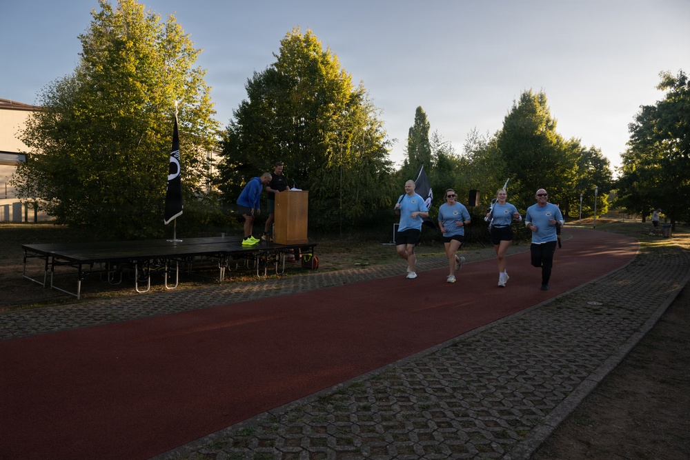 A group of four runners in matching light blue shirts are jogging along a red rubberized track, carrying black flags during what appears to be a commemorative or ceremonial run. A wooden podium and a raised stage platform are set up alongside the track, with two officials or organizers standing nearby. The event takes place outdoors in a park-like setting with tall trees and a building visible in the background during early morning or late afternoon light.