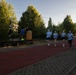 A group of four runners in matching light blue shirts are jogging along a red rubberized track, carrying black flags during what appears to be a commemorative or ceremonial run. A wooden podium and a raised stage platform are set up alongside the track, with two officials or organizers standing nearby. The event takes place outdoors in a park-like setting with tall trees and a building visible in the background during early morning or late afternoon light.