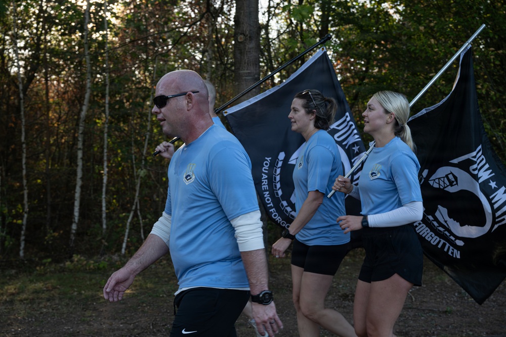 Three individuals wearing matching light blue athletic shirts are walking or marching along an outdoor trail surrounded by trees. Two of them are carrying POW/MIA flags, which are black with the iconic silhouette and the text 'YOU ARE NOT FORGOTTEN.' This appears to be a memorial run or ruck march honoring prisoners of war and those missing in action.