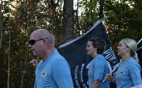 Three individuals wearing matching light blue athletic shirts are walking or marching along an outdoor trail surrounded by trees. Two of them are carrying POW/MIA flags, which are black with the iconic silhouette and the text 'YOU ARE NOT FORGOTTEN.' This appears to be a memorial run or ruck march honoring prisoners of war and those missing in action.