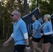 Three individuals wearing matching light blue athletic shirts are walking or marching along an outdoor trail surrounded by trees. Two of them are carrying POW/MIA flags, which are black with the iconic silhouette and the text 'YOU ARE NOT FORGOTTEN.' This appears to be a memorial run or ruck march honoring prisoners of war and those missing in action.