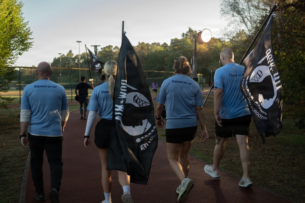 A group of individuals wearing matching light blue shirts with 'Depend on us!' printed on the back are walking along an outdoor track while carrying POW/MIA flags. The scene appears to take place during early morning or evening hours at a recreational sports facility. This appears to be a memorial or awareness walk honoring prisoners of war and those missing in action.
