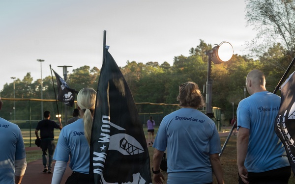 A group of individuals wearing matching light blue shirts with 'Depend on us!' printed on the back are walking along an outdoor track while carrying POW/MIA flags. The scene appears to take place during early morning or evening hours at a recreational sports facility. This appears to be a memorial or awareness walk honoring prisoners of war and those missing in action.