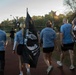 A group of individuals wearing matching light blue shirts with 'Depend on us!' printed on the back are walking along an outdoor track while carrying POW/MIA flags. The scene appears to take place during early morning or evening hours at a recreational sports facility. This appears to be a memorial or awareness walk honoring prisoners of war and those missing in action.