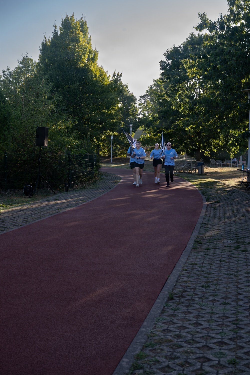 A group of three runners in matching light blue t-shirts and black shorts are jogging along a red rubberized running path in a park setting. Two of the runners are carrying flags or banners, suggesting this may be an organized relay or ceremonial run. The scene is set in a tree-lined park with cobblestone borders flanking the track.
