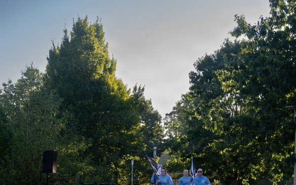 A group of three runners in matching light blue t-shirts and black shorts are jogging along a red rubberized running path in a park setting. Two of the runners are carrying flags or banners, suggesting this may be an organized relay or ceremonial run. The scene is set in a tree-lined park with cobblestone borders flanking the track.