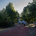 A group of three runners in matching light blue t-shirts and black shorts are jogging along a red rubberized running path in a park setting. Two of the runners are carrying flags or banners, suggesting this may be an organized relay or ceremonial run. The scene is set in a tree-lined park with cobblestone borders flanking the track.