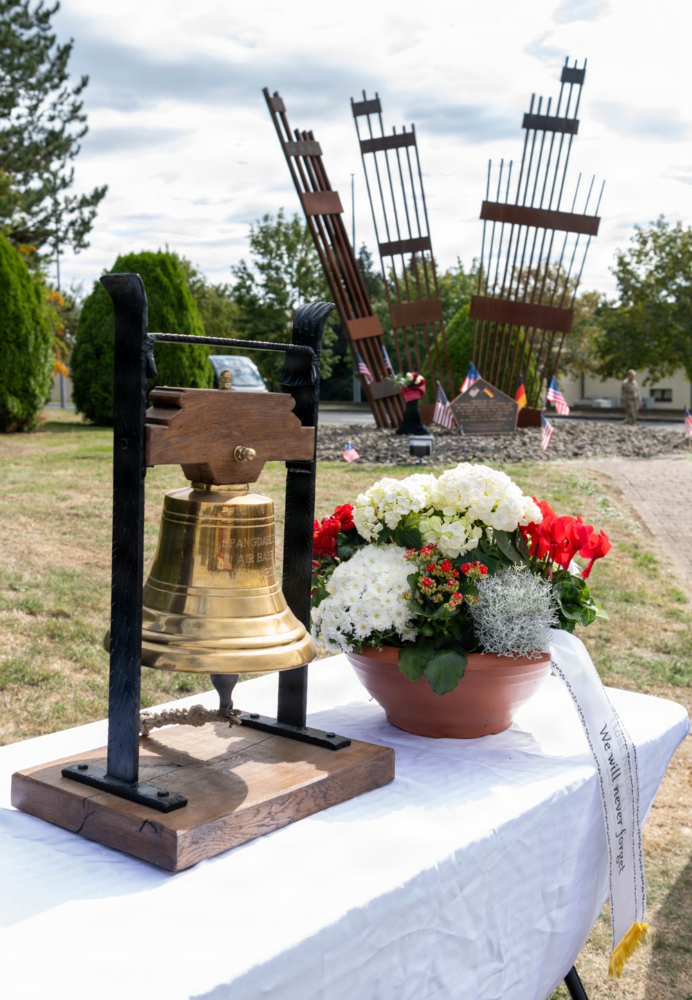 A memorial ceremony setup featuring a brass bell engraved with 'Spangdahlem Air Base' on a wooden and iron stand, placed on a white-draped table alongside a floral arrangement. In the background stands a 9/11 memorial sculpture made of steel beams resembling the collapsed Twin Towers, surrounded by American and German flags. A sash on the table reads 'We will never forget,' indicating this is a September 11 commemoration event.
