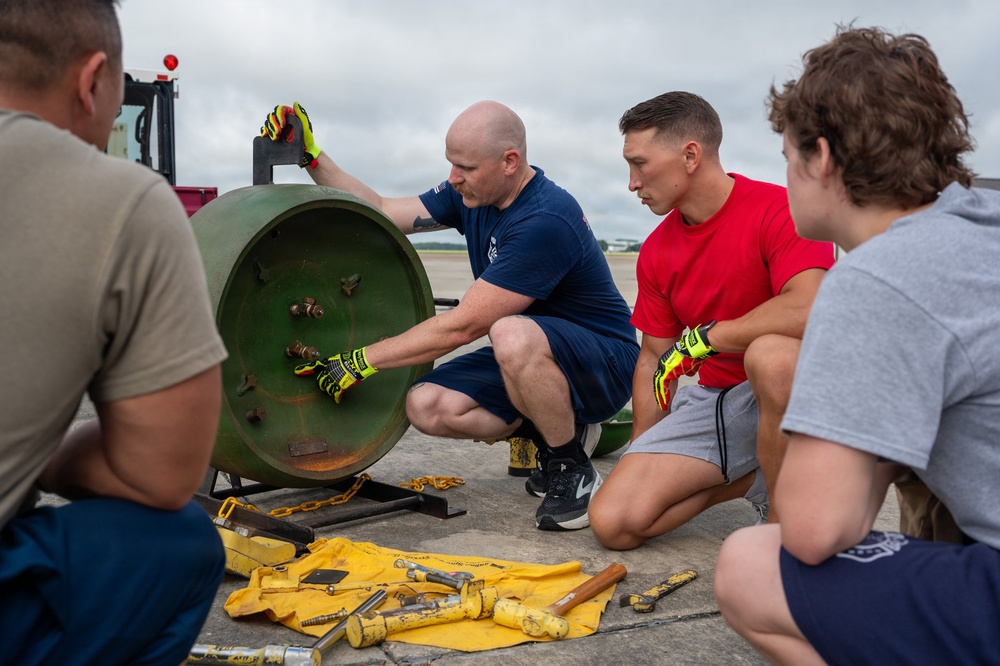 A group of four individuals, appearing to be military or aviation personnel, are gathered around a large green cylindrical object on a tarmac or airfield surface. One bald man wearing a navy blue shirt and yellow safety gloves is demonstrating or instructing others on how to handle or disassemble the cylindrical component. Tools including hammers, bolts, and other hardware are laid out on a yellow cloth on the ground.