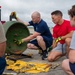 A group of four individuals, appearing to be military or aviation personnel, are gathered around a large green cylindrical object on a tarmac or airfield surface. One bald man wearing a navy blue shirt and yellow safety gloves is demonstrating or instructing others on how to handle or disassemble the cylindrical component. Tools including hammers, bolts, and other hardware are laid out on a yellow cloth on the ground.